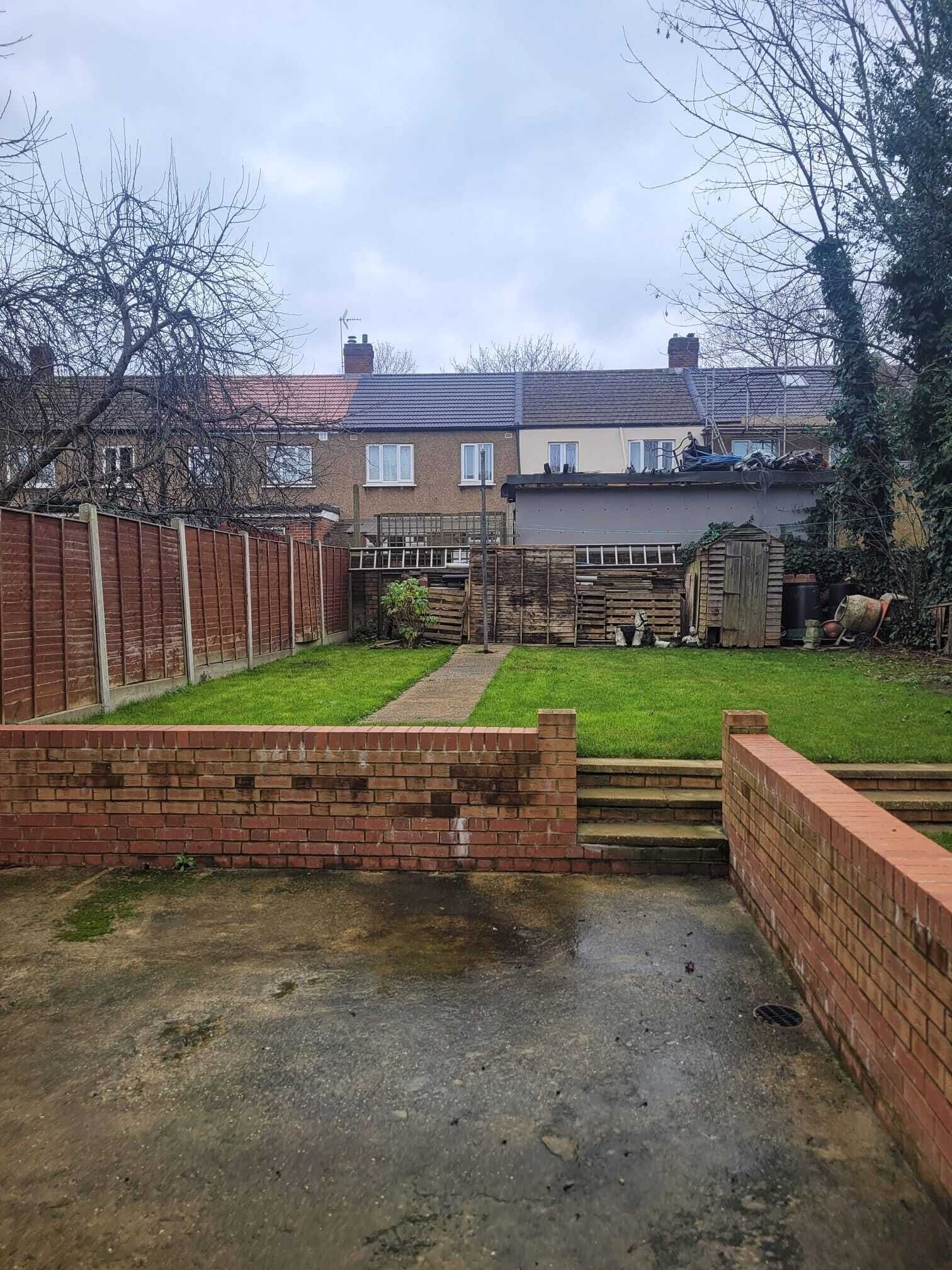A residential backyard with a brick wall, a grassy lawn, and a concrete path leading to a house.