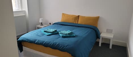 Un lit bien fait, avec une literie bleue et jaune, deux tables de chevet blanches, une lampe et une fenêtre avec des stores.