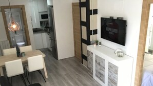 Una sala moderna con un sofá negro, una zona de comedor con una mesa de madera y sillas blancas, una cocina con armarios blancos, y un televisor montado en la pared.