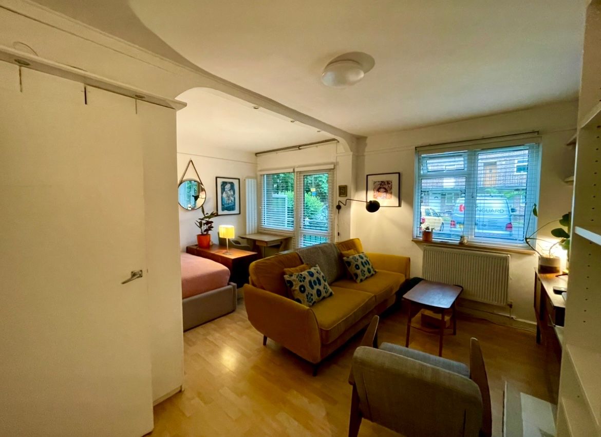 A compact living space with a sofa, a small table, and a chair. There is a window with blinds, a radiator, and a door leading to another room.