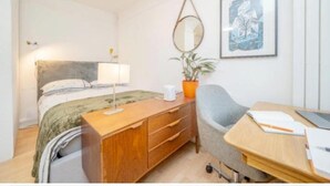 Een slaapkamer met een bed, nachtkastje, lamp, stoel, bureau en een lijst aan de muur.