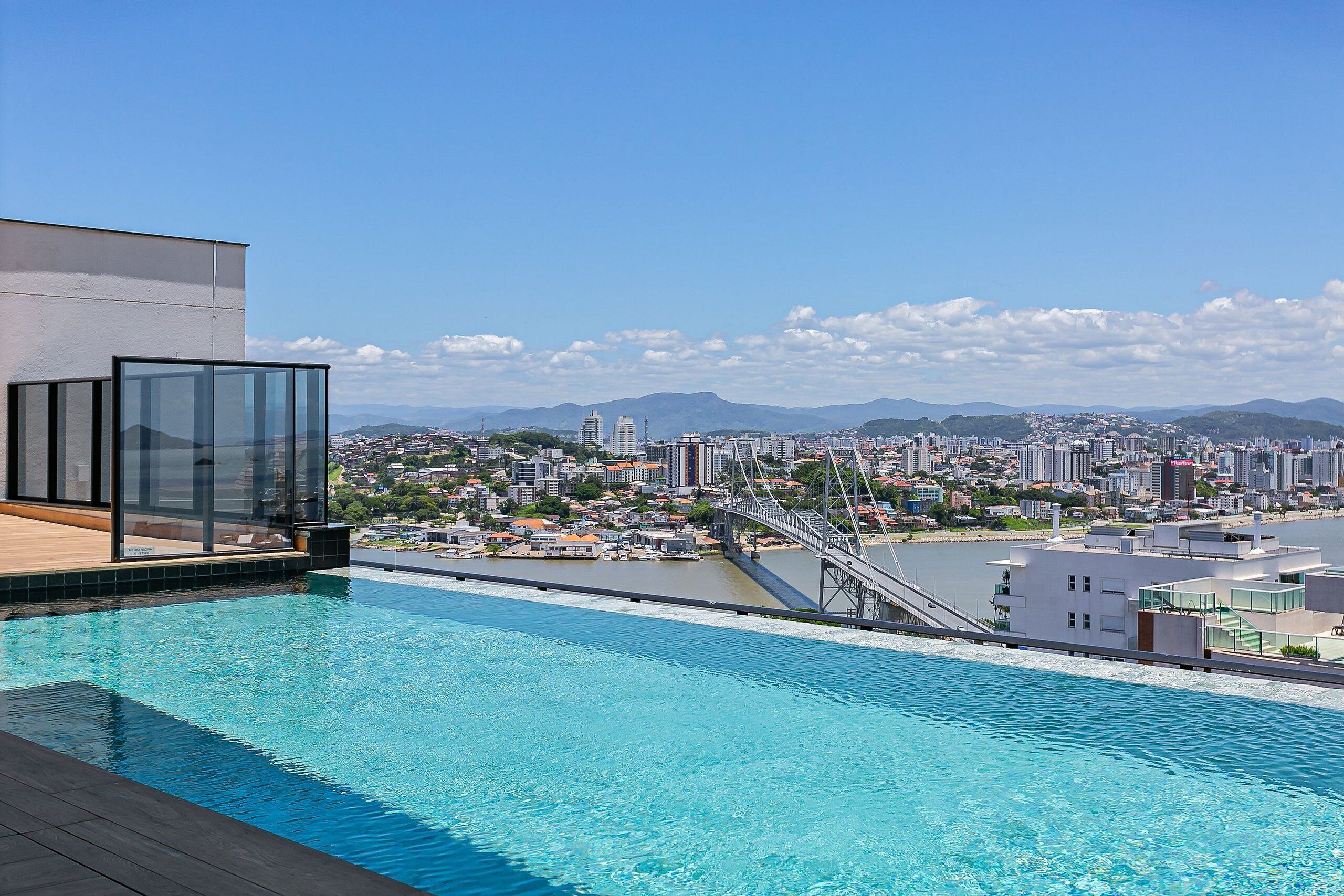 Una piscina en la azotea con vistas a la ciudad, un salón con paredes de vidrio y un puente al fondo.