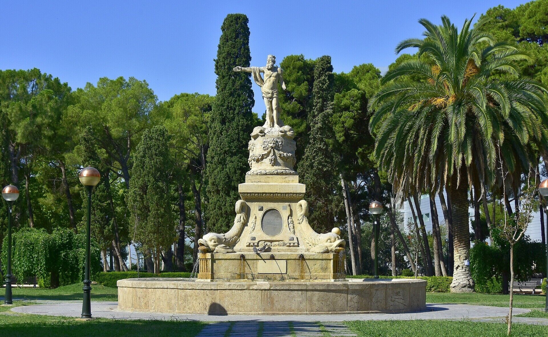 A classical statue on a pedestal in a park with palm trees and a clear sky.