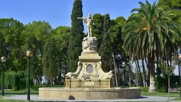 A classical statue on a pedestal in a park with palm trees and a clear sky.