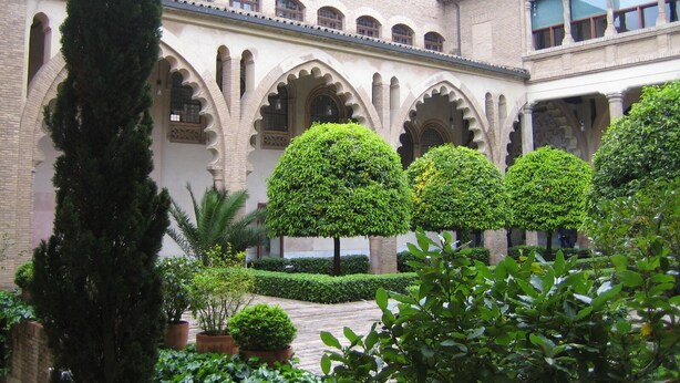 Un patio con arcos, árboles en macetas y un edificio de ladrillo con varias ventanas.