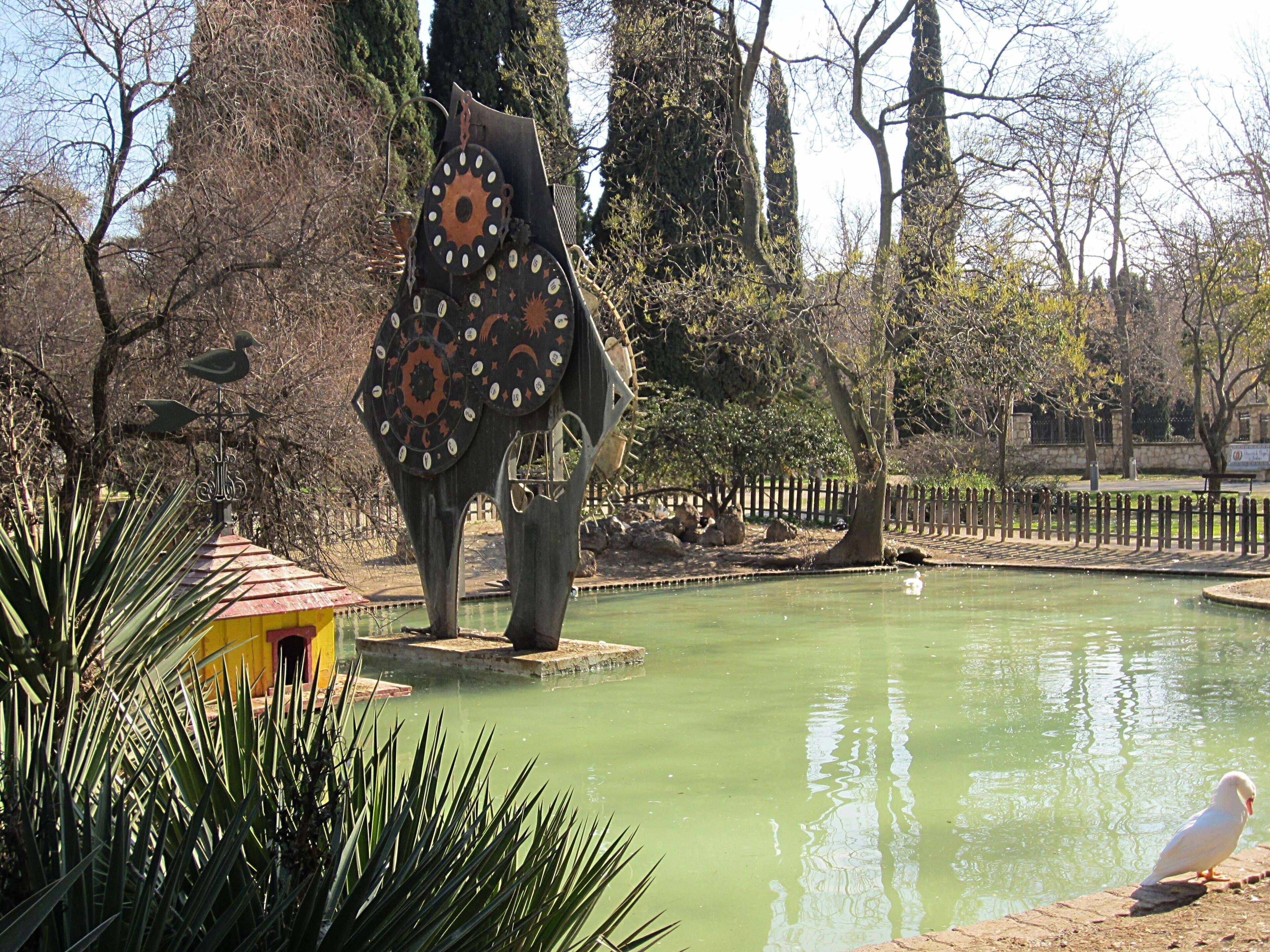 Una escultura de una figura con forma de ave, con un diseño circular en el cuerpo, ubicada sobre una plataforma en un parque con un estanque y árboles de fondo.