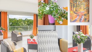 A balcony with a view of greenery, a wicker sofa, a table with a vase of flowers, and a painting on the wall.