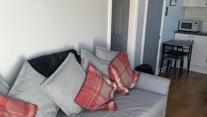 A grey sofa with red and beige pillows, a framed picture on the wall, and a kitchen area with a microwave and a small table. - Modern One Bedroom Chalet With Central Heating (Bridlington)