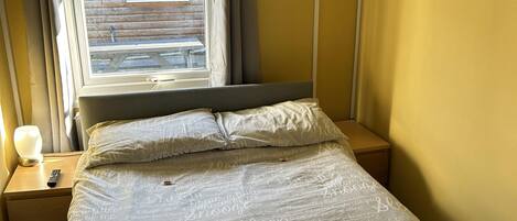 Un dormitorio con una cama, una mesita de noche, una lámpara, una ventana con cortinas y vistas al exterior.