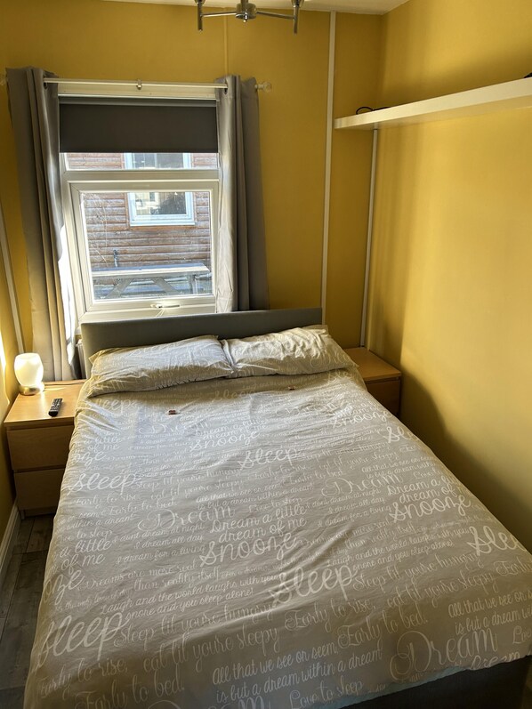 A bedroom with a bed, a nightstand, a lamp, a window with curtains, and a view of the outside. - Modern One Bedroom Chalet With Central Heating (Bridlington)