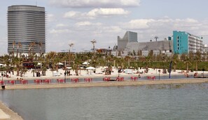 Küstenlandschaft mit Strand, Palmen und einem Wasserabschnitt. Rote Stühle und Sonnenschirme sind vorhanden, im Hintergrund ein hohes Gebäude.
