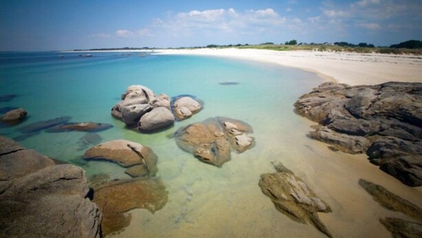A beach with clear turquoise water, large rocks in the foreground, and a sandy beach extending to the horizon.
