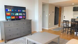 Un salon moderne avec un téléviseur à écran plat, une commode grise et un coin repas comprenant une table et des chaises.
