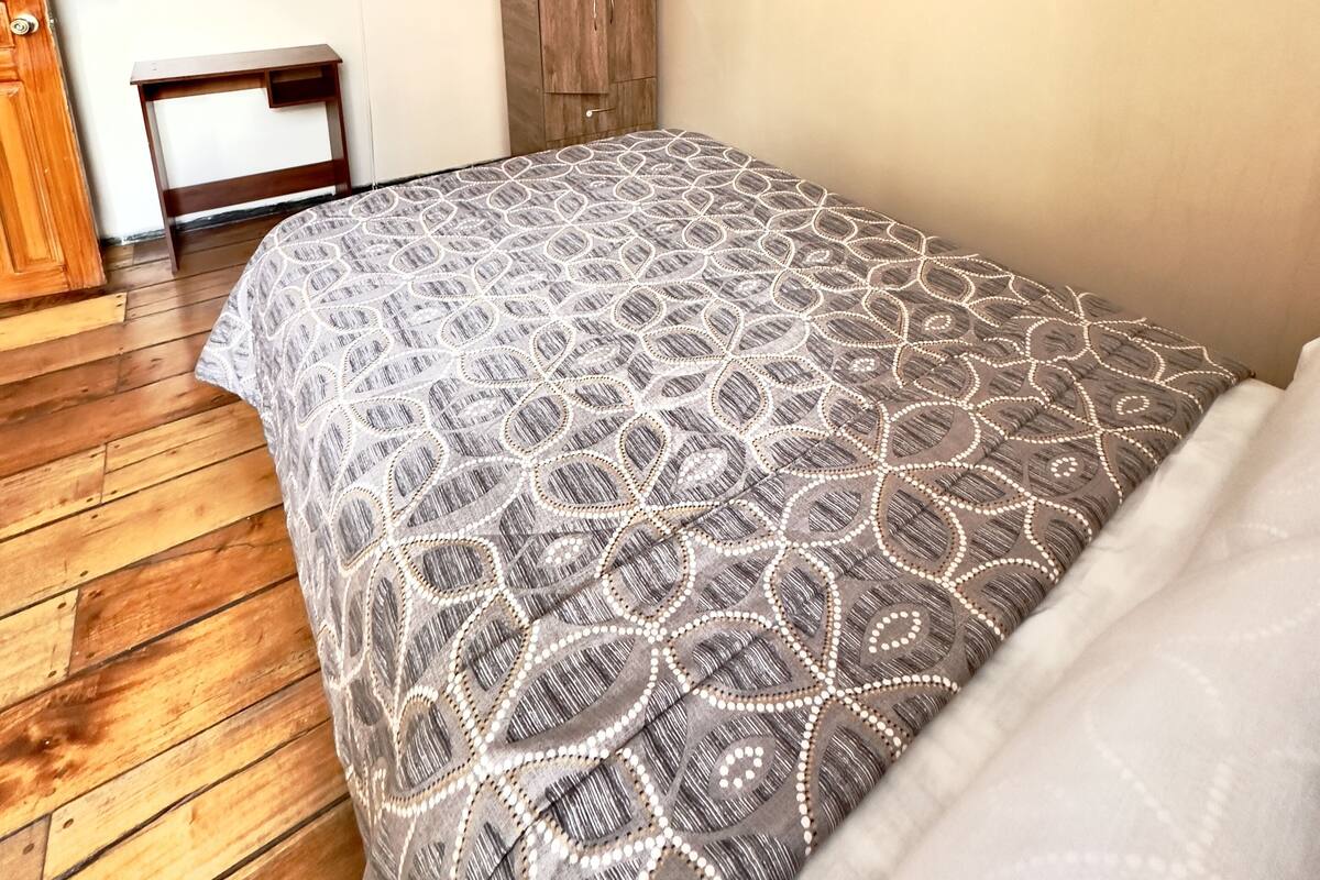 A bedroom with a patterned bedspread, a wooden wardrobe, a television mounted on the wall, and a wooden floor.