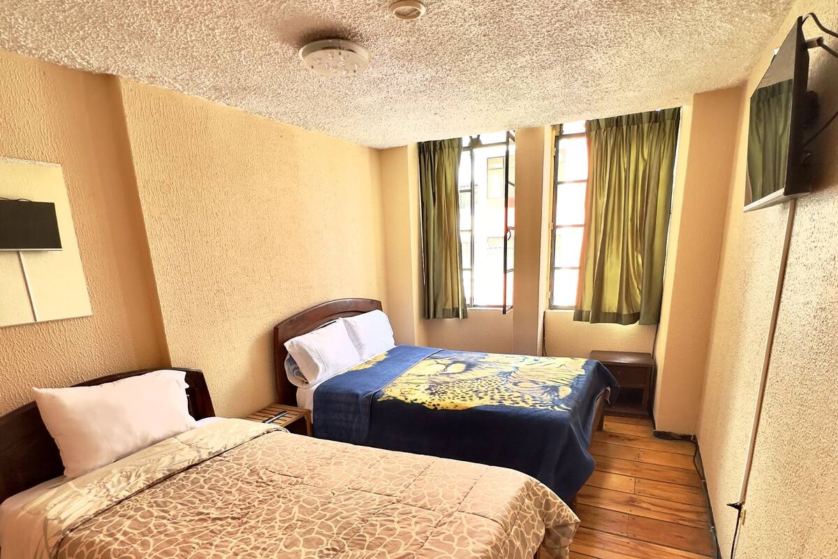 A hotel room with two beds, wooden flooring, a ceiling with a textured finish, and a window with green curtains.