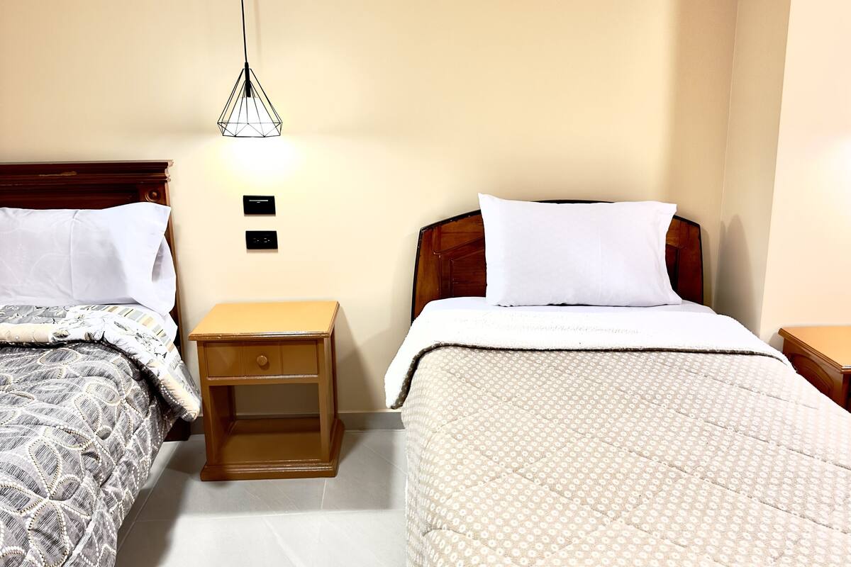 A hotel room with two beds, a nightstand, a wall-mounted light, and a wooden headboard.