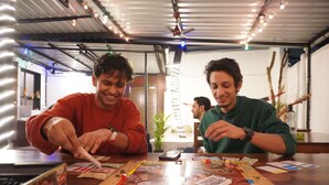 Dos personas jugando un juego de mesa en una mesa, con un ventilador de techo, luces colgantes y una planta al fondo.