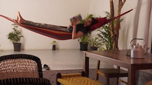 Una persona recostada en una hamaca dentro de una habitación con una mesa de madera, sillas y plantas en macetas.