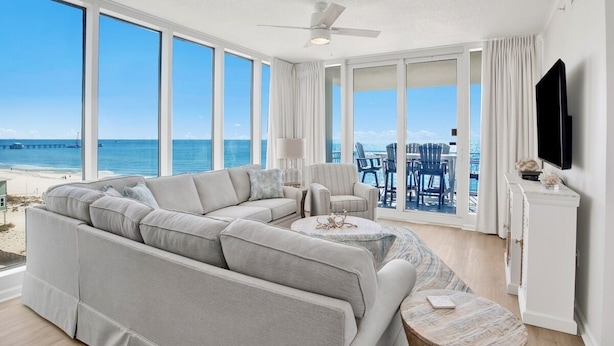 A spacious living room with a large sectional sofa, a small round coffee table, and a ceiling fan. There is a large window with a view of the beach and ocean. - Colonnades 601 | Beach Front Luxury in Gulf Shores (Gulf Shores)