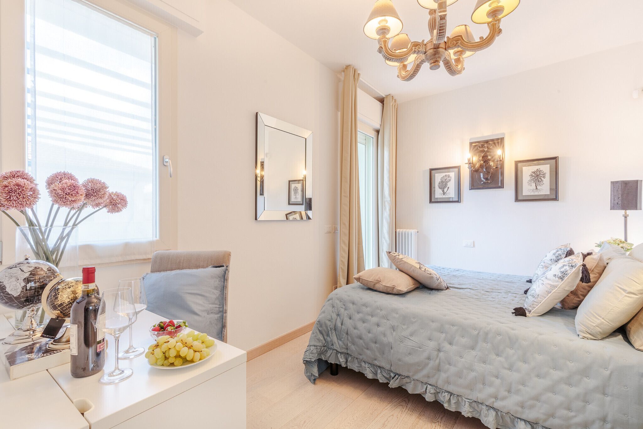 Un dormitorio con una cama, una mesita de noche con una botella, vasos y uvas, un globo terráqueo y una ventana con cortinas.