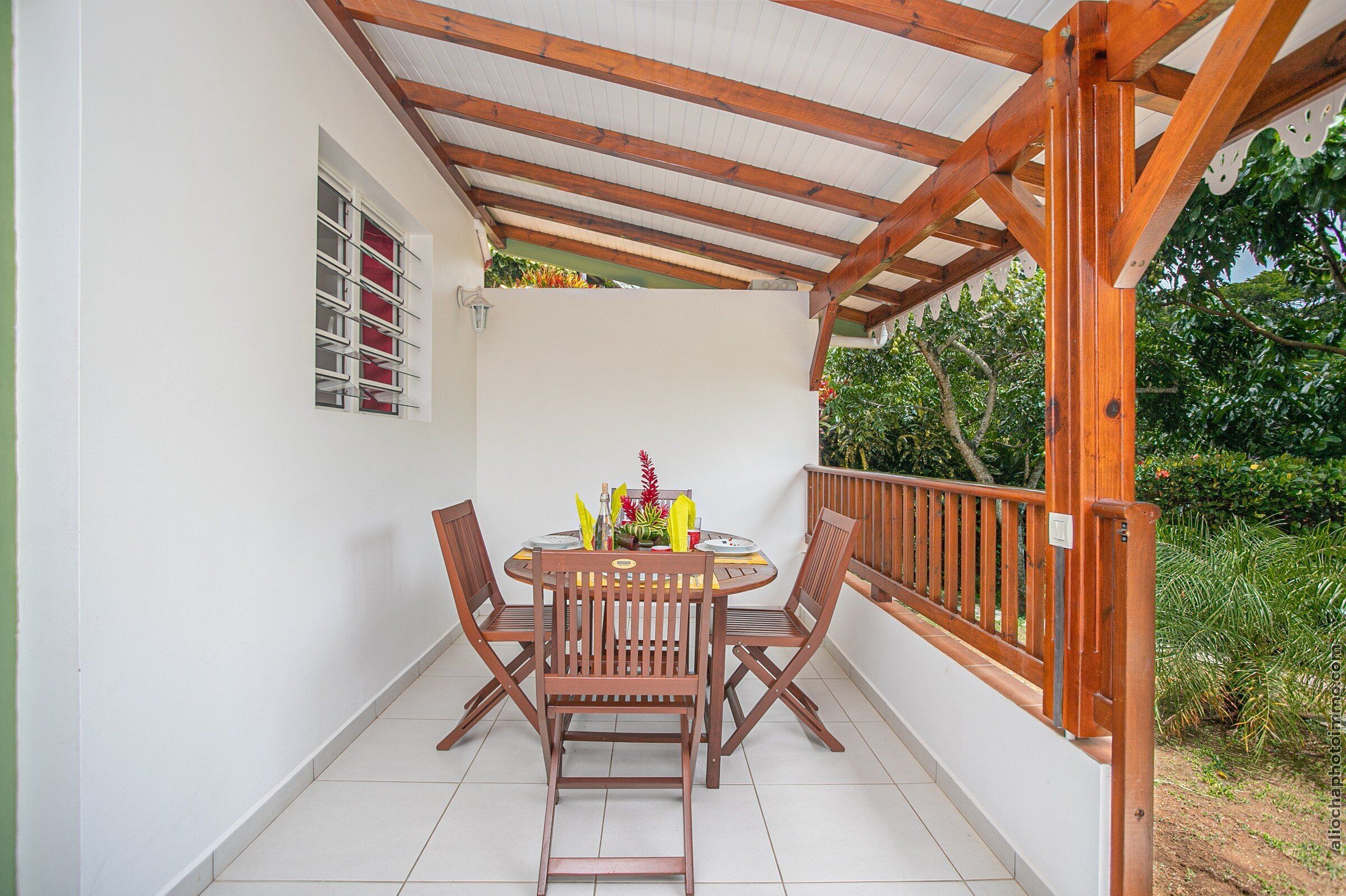 A covered patio with a wooden table and chairs, a small window with red curtains, and a view of greenery.