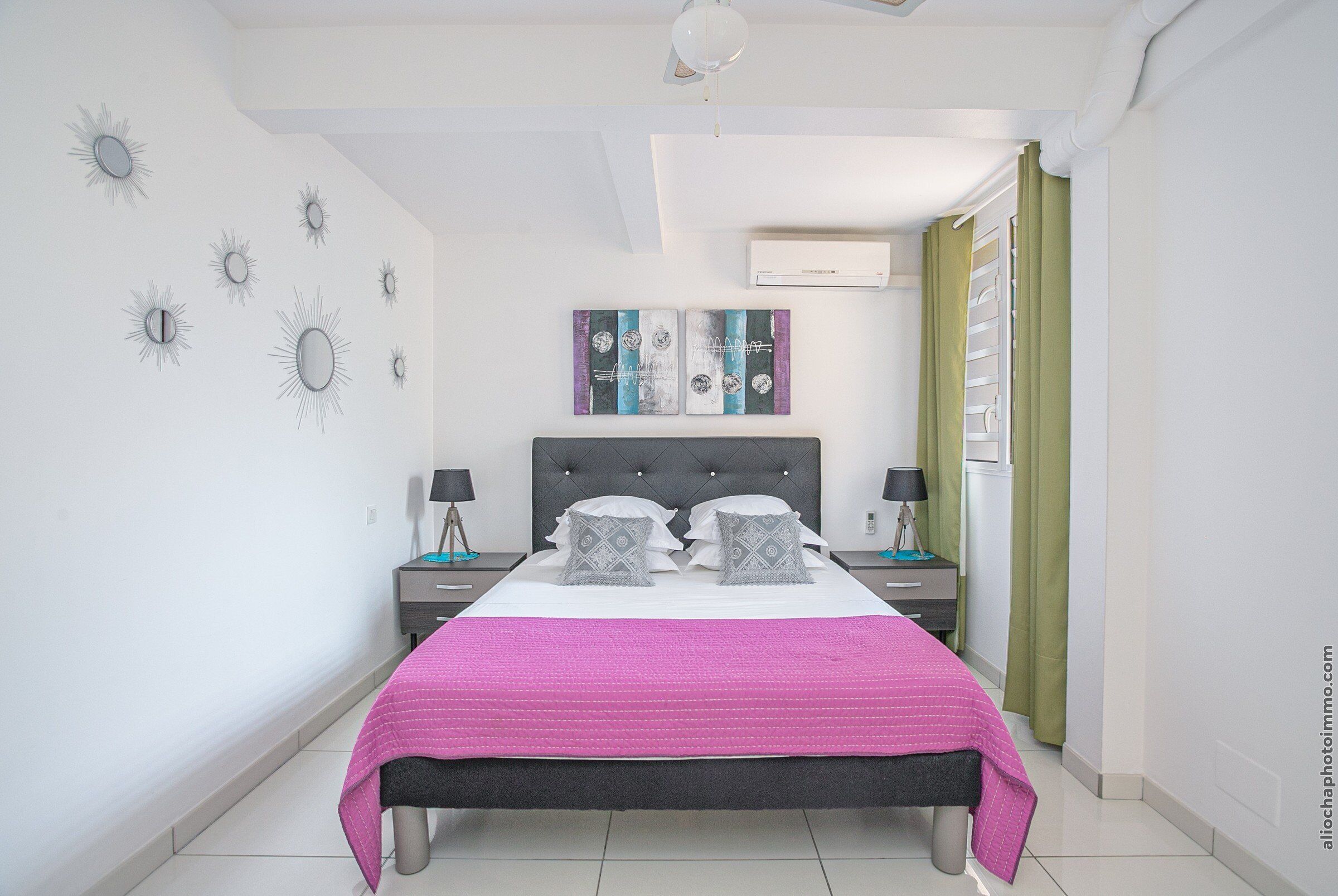 Une chambre à coucher comprenant un lit, deux tables de chevet, un ventilateur de plafond, des œuvres d’art murales et un appareil de climatisation.