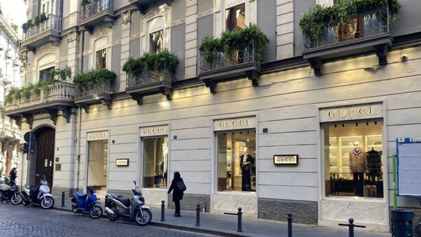 En byggnad med flera butikslokaler, inklusive en framträdande Gucci-butik. Det står parkerade motorcyklar och en person går på trottoaren.
