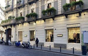 Ein Gebäude mit mehreren Schaufenstern, darunter ein gut sichtbarer Gucci-Store. Auf dem Bürgersteig stehen Roller und eine Fußgängerin.