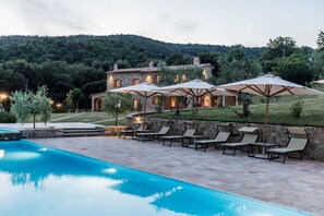 A pool area with lounge chairs and umbrellas, a stone building with multiple windows, and a hillside in the background.