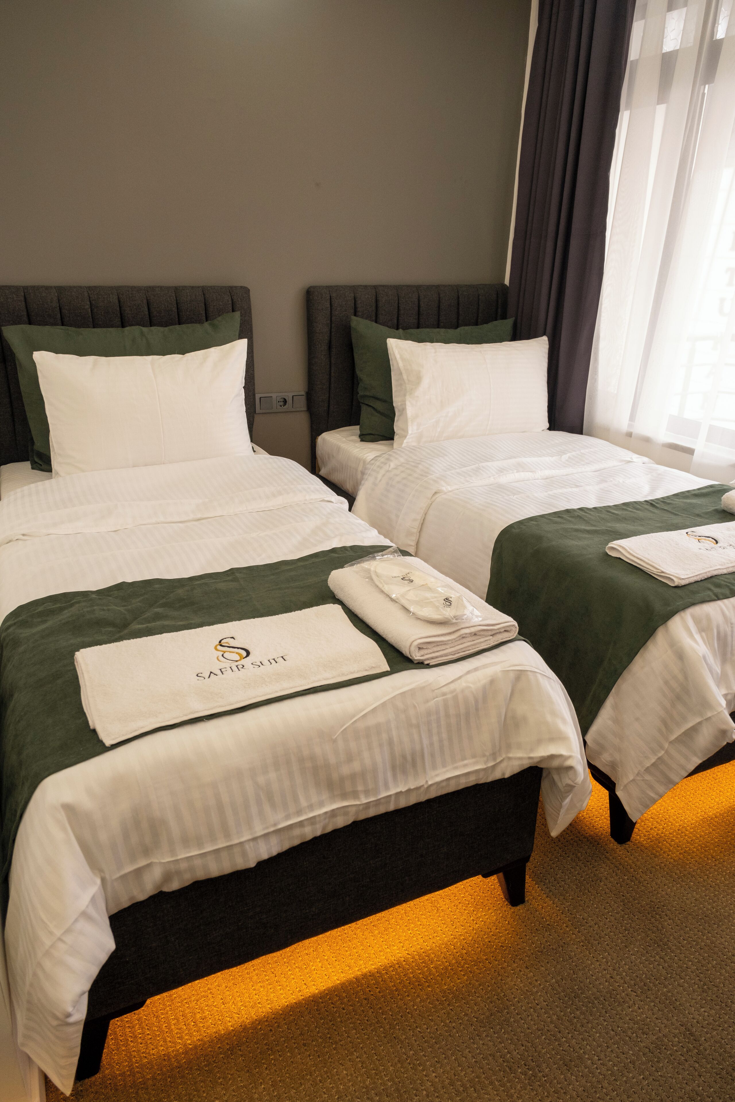 A hotel room with two beds, each with a green bedspread and white linens. There are towels with the hotel's logo on the beds.
