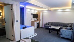 A modern hotel room with a sofa, a small table, a TV, a minibar, and a bathroom with a bathtub.