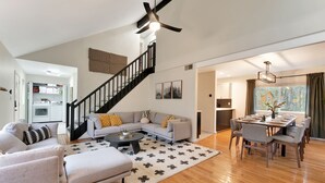 A modern living room with a gray sofa set, a coffee table, and a dining area with a table and chairs. A black staircase leads to an upper level. - Poconos FUN! Hot Tub, Fire Pit, Game Room, Arcade (East Stroudsburg)