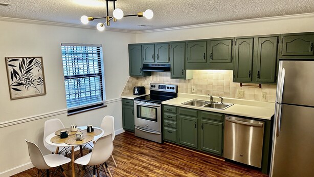 Une cuisine moderne avec des meubles de rangement verts, des appareils en acier inoxydable et un coin repas comprenant une table et des chaises blanches.