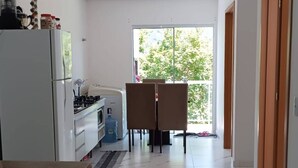 Una cocina moderna con refrigerador, estufa y comedor con sillas. Hay un sofá en primer plano y se ve follaje verde al exterior.