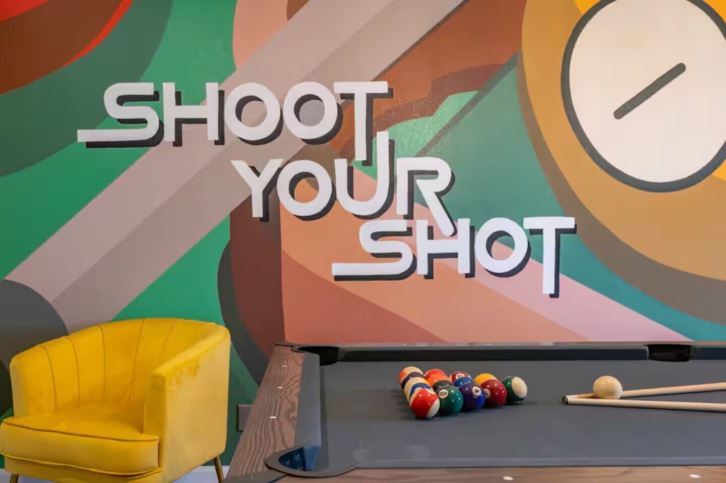 Una mesa de billar con bolas y tacos, una silla amarilla y una pared con el texto "SHOOT YOUR SHOT".
