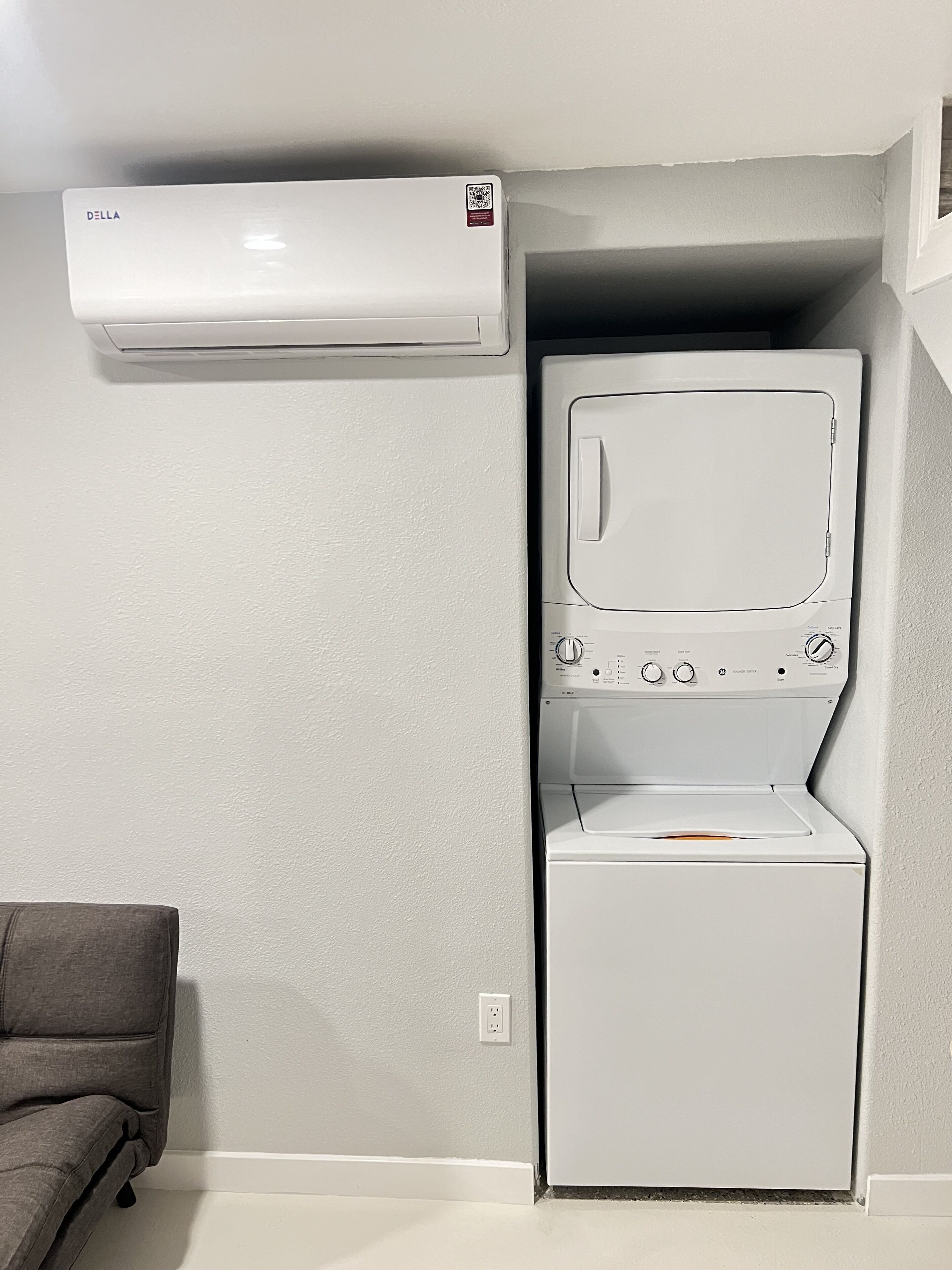 Ein kleiner Waschraum mit einer Frontlader-Waschmaschine und einem Trockner, einer Wand-Klimaanlage und einem grauen Sofa.