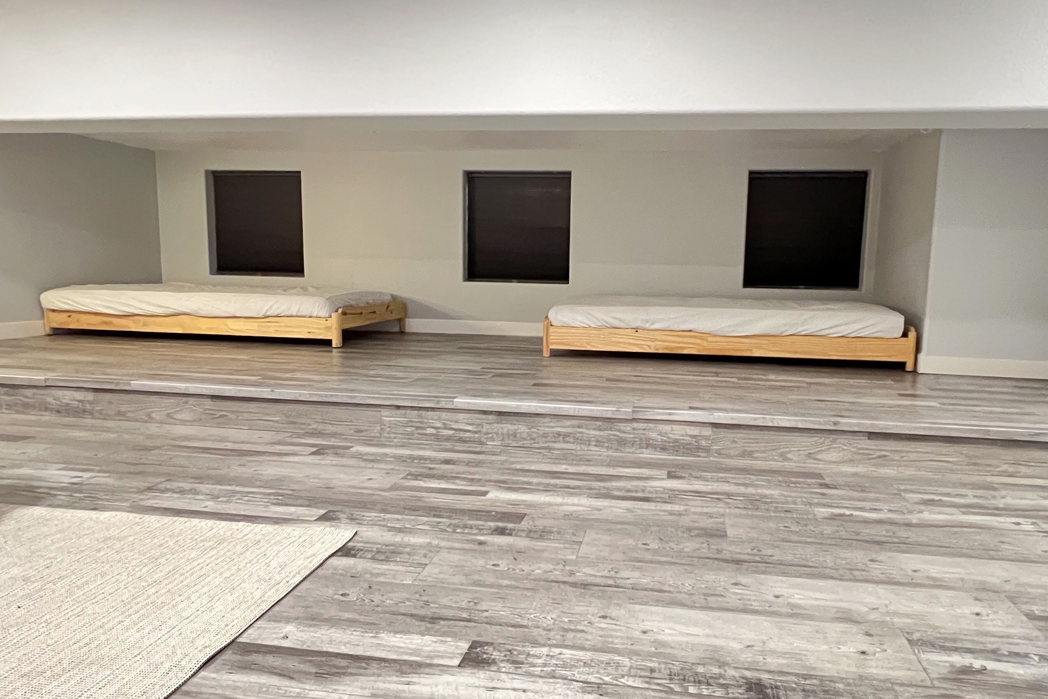 Una habitación con dos camas de madera, piso gris y tres ventanas pequeñas.