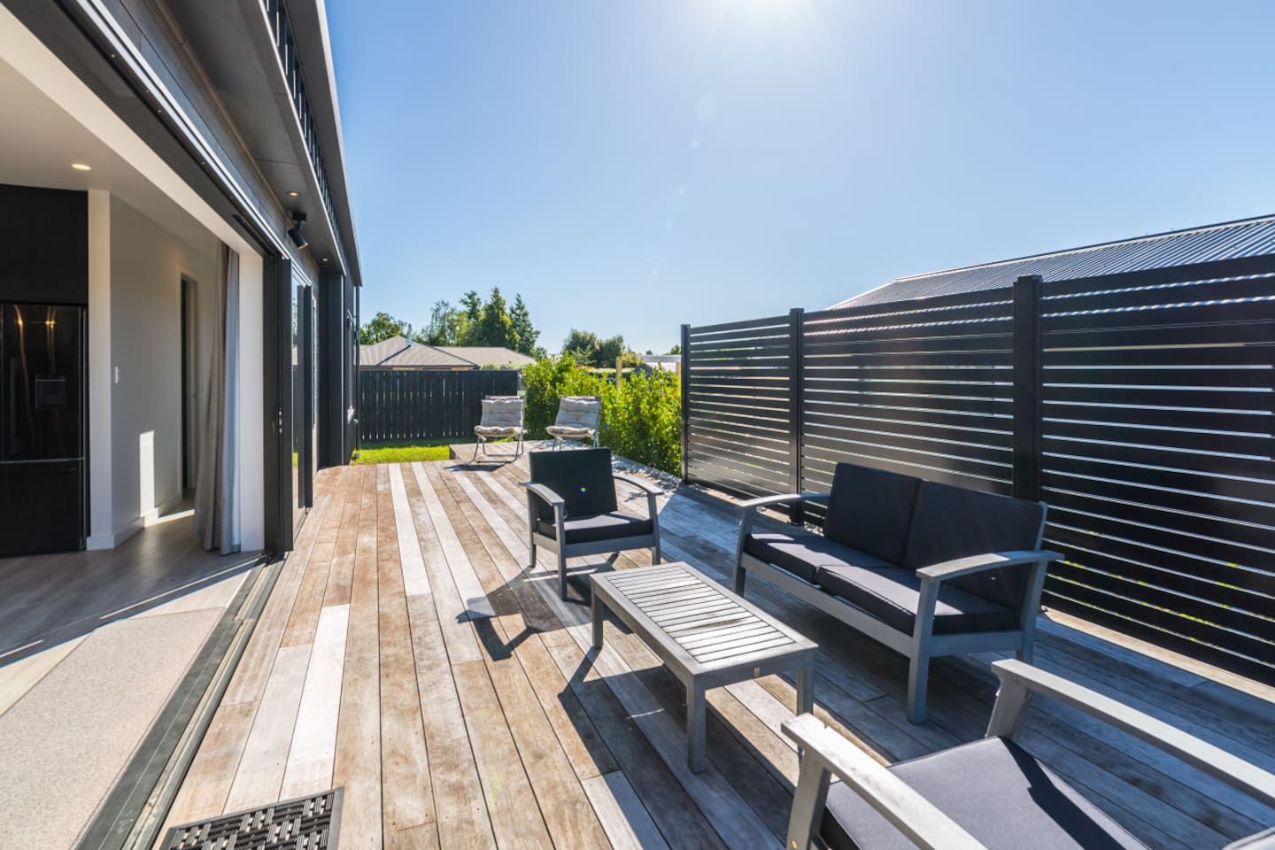 Een terras buiten met houten vloer, een bank, twee stoelen en een bijzettafeltje.