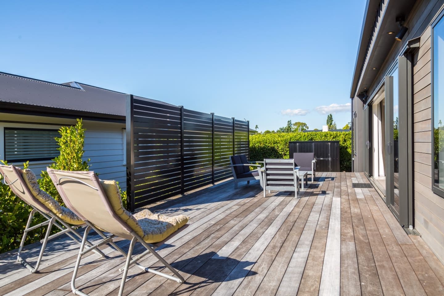 Une terrasse en bois avec des meubles d’extérieur, une clôture assurant l’intimité et un bâtiment doté de grandes fenêtres.