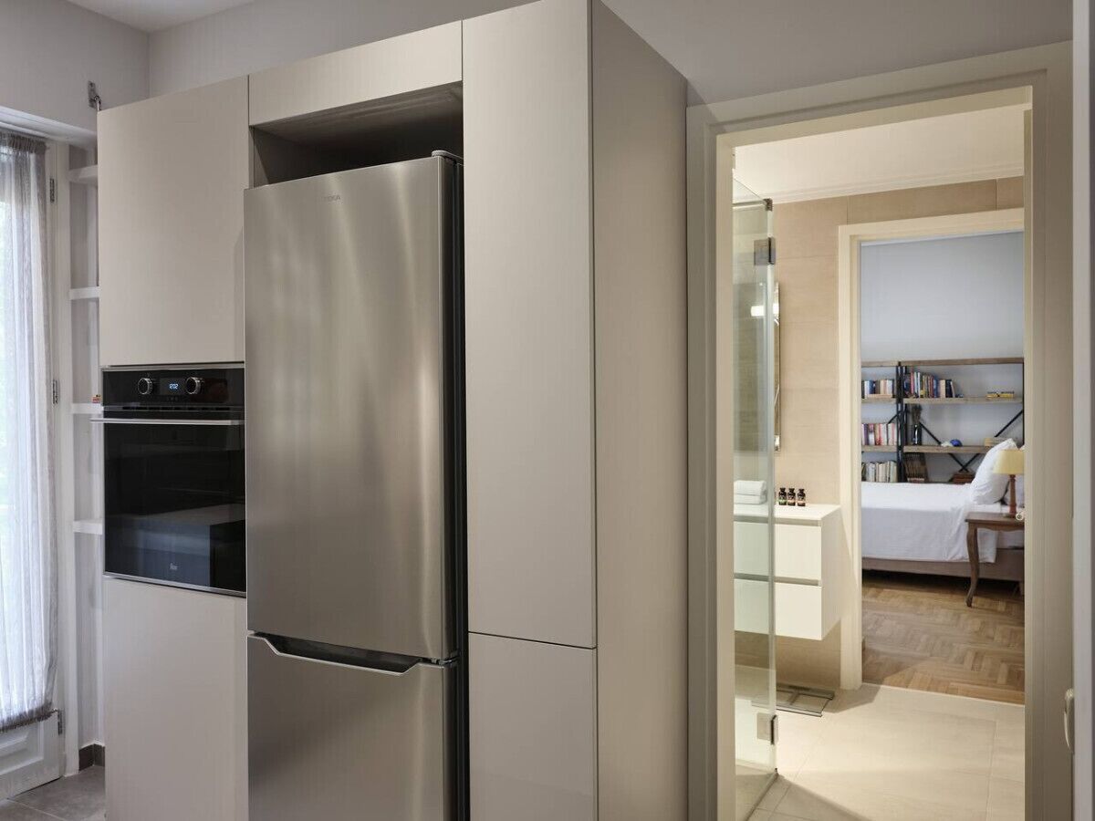 A modern kitchen with a stainless steel refrigerator, built-in oven, and a glass door leading to a bedroom with a bed and a bedside table.