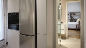 A modern kitchen with a stainless steel refrigerator, built-in oven, and a glass door leading to a bedroom with a bed and a bedside table. - Elegant Boutique Apt in Kifissia (Kifisia)