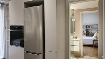 A modern kitchen with a stainless steel refrigerator, built-in oven, and a glass door leading to a bedroom with a bed and a bedside table.