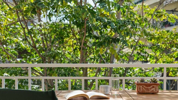A wooden table with an open book, a glass of water, and a potted plant on a balcony with a white railing. - Elegant Boutique Apt in Kifissia (Kifisia)