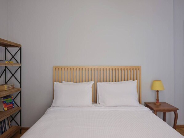 Una cama bien hecha con cabecera de madera, una mesita de noche con lámpara y una estantería con libros.