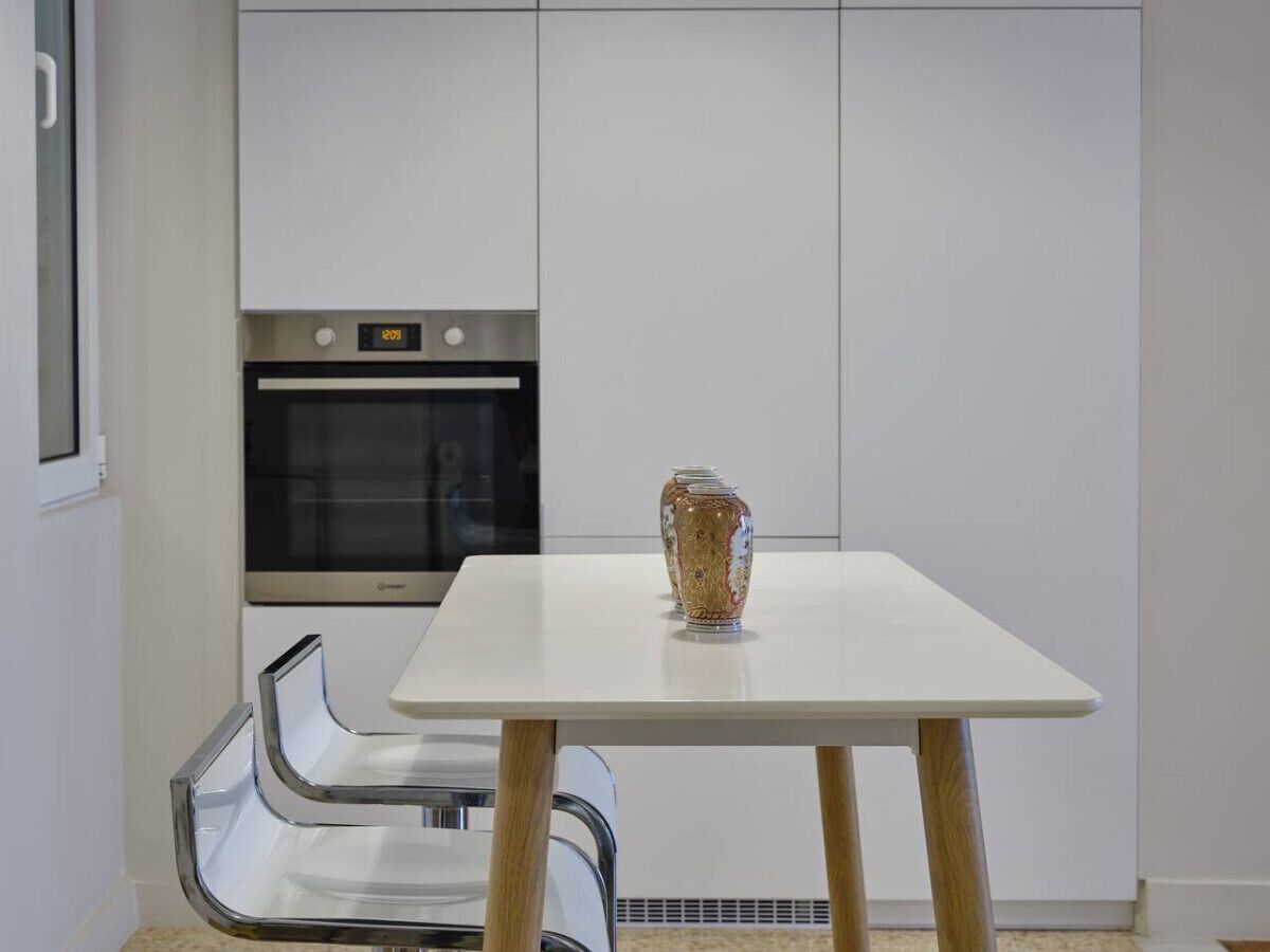 Eine moderne Küche mit weißen Schränken, einem Einbauofen, einem weißen Tisch mit einer dekorativen Vase und einem Glastischstuhl.