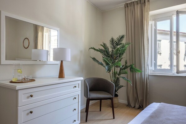 Une chambre d’hôtel comprenant un lit, une commode avec des tiroirs, une chaise, une plante en pot, une lampe et une fenêtre avec des rideaux.