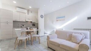 Eine moderne Küche mit weißen Schränken, einem Esstisch mit Stühlen und einem beige gefärbten Sofa in einem hellen Wohnbereich mit Fliesenboden.
