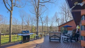 A wooden deck with a grill, picnic table, and chairs, overlooking a grassy area with trees.