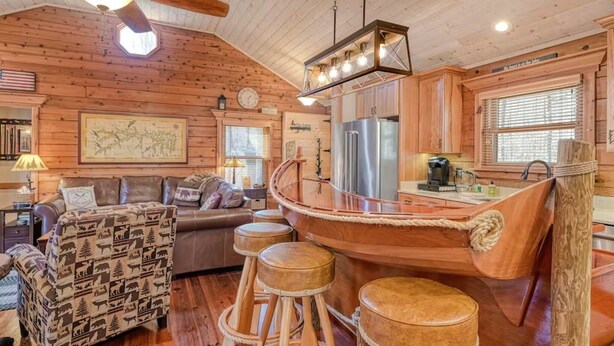 A cabin interior with a wooden boat-shaped bar, a sofa with animal print cushions, a wooden ceiling fan, and a map on the wall.