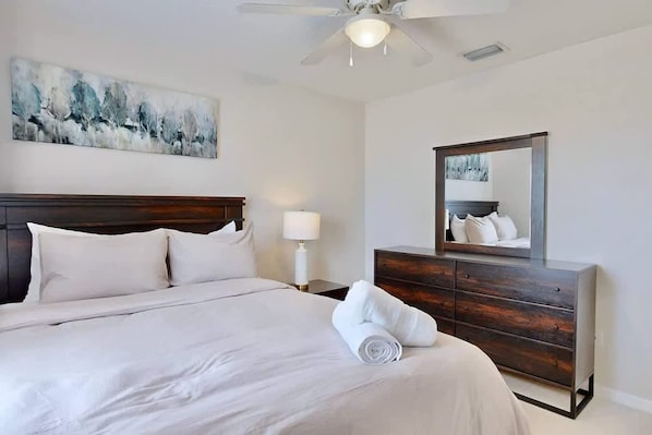 Una habitación de hotel con una cama, una mesita de noche, una lámpara, un aparador, un ventilador de techo y un cuadro en la pared.